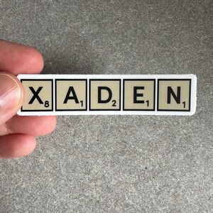 May include: A rectangular sticker with the name "XADEN" spelled out using Scrabble tiles. The tiles are beige with black letters and numbers, and are arranged in a single row on a white background.