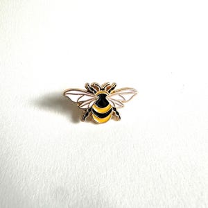 May include: A gold-toned bee pin with black, yellow, and white enamel detailing. The bee has outstretched wings and a detailed body. This decorative pin is a charming accessory for jackets, bags, or hats.
