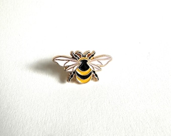 May include: A gold-toned bee pin with black, yellow, and white enamel detailing. The bee has outstretched wings and a detailed body. This decorative pin is a charming accessory for jackets, bags, or hats.