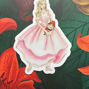 May include: A sticker featuring a watercolor illustration of a woman in a pink ball gown holding a nutcracker. The woman has long blonde hair and is wearing a necklace. The sticker has a white border and is set against a floral background.