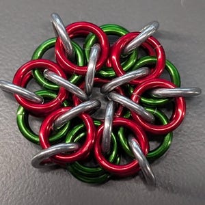 May include: A handmade chainmaille flower design. The flower is composed of red and green anodized aluminum rings, with silver-colored rings connecting them. The piece is displayed on a gray surface.