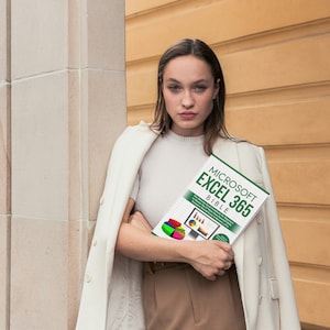 A lady wearing a long cream coat draped over both her shoulders is staring straight in front. She is leaning against a stone pillar holding a Microsoft Excel book called Microsoft Excel 365 Bible which is authored by Excel expert Harjit Suman.