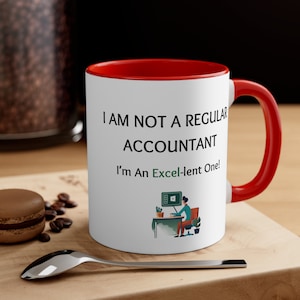 Könnte beinhalten: Eine weiße Kaffeetasse mit rotem Rand und Henkel. Die Tasse hat einen schwarzen Text mit der Aufschrift "Ich bin kein normaler Buchhalter, ich bin ein Excel-lent!" und eine Cartoon-Illustration einer Person, die an einem Computer arbeitet.