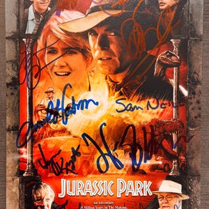 May include: A rectangular Jurassic Park movie poster featuring a collage of cast members and a dinosaur, with the text "Life Finds A Way" and "Jurassic Park An Adventure 65 Million Years In The Making". The poster is covered in multiple ink signatures.