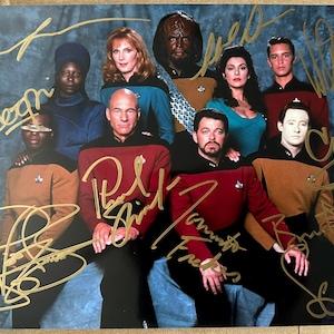 May include: A group photo of the cast of Star Trek: The Next Generation, featuring the actors in their Starfleet uniforms. The photo is signed in gold ink by the cast members. The background is a textured blue surface.