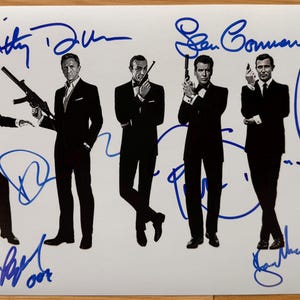 May include: A white print featuring six men in black suits, each holding a gun or weapon, with blue signatures above and below. The image is a promotional item or collectible.