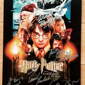 May include: A movie poster for the film Harry Potter and the Sorcerer's Stone. The poster features a dark background with a large image of Harry Potter in the center. The poster also features images of other characters from the film, including Ron Weasley, Hermione Granger, and Hagrid. The poster is signed by the cast members, including Daniel Radcliffe, Rupert Grint, Emma Watson, and Robbie Coltrane.