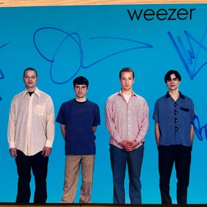 May include: A signed Weezer band photo featuring four men in casual clothing against a blue background. The band members are Rivers Cuomo, Pat Wilson, Brian Bell, and Scott Shriner. The photo is signed by all four band members in blue ink.