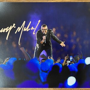 May include: A black and white photo of a man singing on stage in front of a crowd. The man is wearing a black jacket and black pants. The crowd is holding up their hands and phones. The man's signature, "George Michael", is written in gold in the top left corner of the photo.