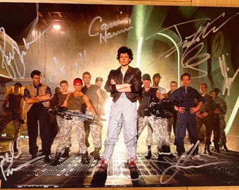 May include: A signed photo of the cast of the movie Aliens, featuring Sigourney Weaver as Ripley, in a futuristic setting. The photo is signed by the actors, including Sigourney Weaver, Carrie Henn, Michael Biehn, Paul Reiser, Lance Henriksen, and others.