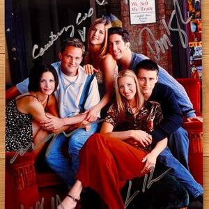 Peut inclure: Photographie couleur dédicacée du casting de la série télévisée Friends. Les acteurs sont assis sur un canapé rouge. En arrière-plan, un mur de briques et une enseigne "We Buy, We Sell, We Trade".
