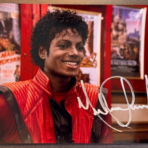 May include: A glossy photograph of Michael Jackson wearing a red jacket with black leather accents, featuring a white ink autograph across the lower right side and a small rectangular sticker in the bottom left corner with the text SERIAL 7858110.