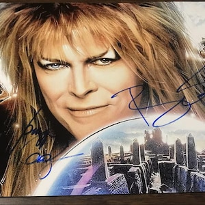 May include: A signed print featuring a portrait of David Bowie from the movie Labyrinth. The image includes characters from the film, with Bowie's face prominently displayed. The print has blue signatures.