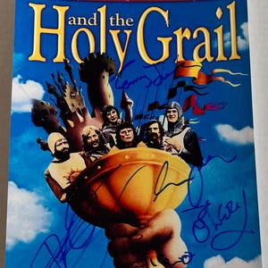 May include: A vintage movie poster for Monty Python and the Holy Grail, featuring the cast members and a blue sky background. The poster is signed by the cast members in blue ink.