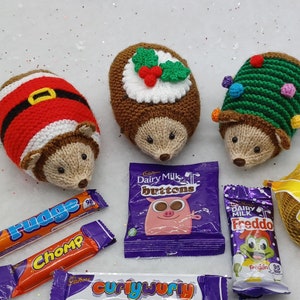 May include: Three knitted hedgehog ornaments, one dressed as Santa, one with a Christmas pudding design, and one with a Christmas tree design.  The ornaments are surrounded by various chocolate bars and a bag of chocolate coins.