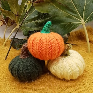 May include: Three knitted pumpkins in orange, dark green, and cream. The orange pumpkin has a green stem, the others have brown and cream stems. They sit on a yellow textured surface, with green leaves behind. A seasonal autumn decoration.