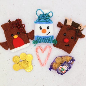 May include: Three knitted Christmas stocking ornaments. A red and brown robin, a white snowman with a blue hat, and a brown reindeer with a red nose. The ornaments are on a white background with a candy cane and chocolate coins.