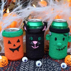 May include: Three Halloween-themed can coolers. One is orange with a jack-o'-lantern face, one is black with a cat face, and the last is green with a Frankenstein face. The coolers are knitted and sit on a patterned surface.