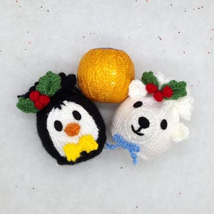 May include: A knitted penguin and polar bear Christmas ornament with holly accents and a gold-wrapped chocolate ball. The penguin has a yellow bow tie, and the polar bear has a blue bow. Festive holiday decor.