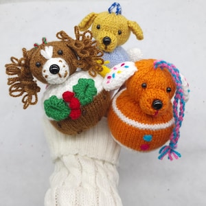 May include: Three handmade knitted dog ornaments featuring a brown spaniel with holly, a yellow dog in a blue jumper, and an orange dog with colourful sprinkles. Each plush toy has black bead eyes and a hanging loop for display on a tree.