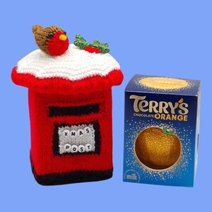 May include: A red knitted post box cover with a white snow-like top, a brown robin bird, and green holly leaves. The front features a grey panel with the text XMAS POST in white beads. Beside it is a blue box of Terry's Chocolate Orange.