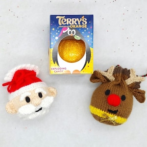 Peut inclure: Une composition festive avec une boîte de Terry's Chocolate Orange, un visage de Père Noël en tricot et une pochette de renne en tricot. La boîte de chocolat présente un design de pingouin. Fond blanc.