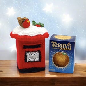 May include: A red knitted post box cover featuring a white snow-like top, a brown robin, and green holly leaves. The front displays a grey square with the text XMAS POST in white beads. Beside it sits a blue box of Terry's Chocolate Orange.