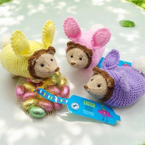 May include: Three knitted hedgehog toys in yellow, pink, and purple, each with bunny ears. A net of foil-wrapped Easter eggs is next to the toys, with a blue tag reading "Easter Eggs". The toys have brown accents and white pom-poms.