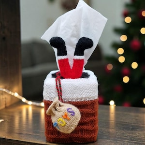 May include: A knitted tissue box cover shaped like a brick chimney with a white top edge. Two red and black legs extend from the top, with a white tissue protruding. A small beige knitted sack labeled TOYS hangs from the side by a red and green cord.