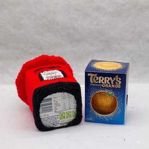 May include: A red and black crocheted cozy shaped like a British post box with the word POST on the front, positioned next to a blue rectangular box of Terry's Chocolate Orange milk chocolate, which features a graphic of a textured orange.