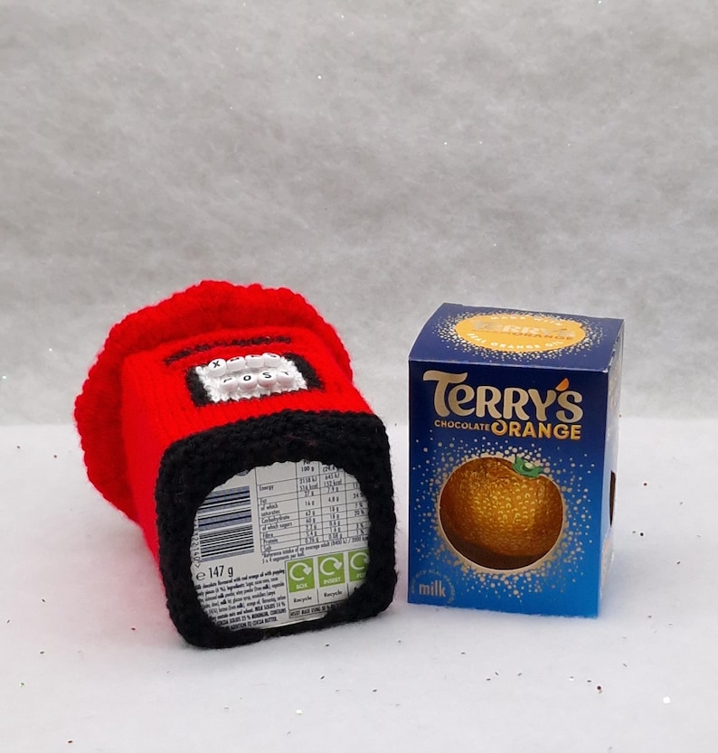 May include: A red and black crocheted cozy shaped like a British post box with the word POST on the front, positioned next to a blue rectangular box of Terry's Chocolate Orange milk chocolate, which features a graphic of a textured orange.