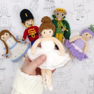 May include: A collection of five handmade knitted dolls featuring a ballerina in a white tutu, a soldier in a red uniform, a mouse in a green cape, a figure in a blue and white dress, and a fairy in a purple dress with wings.