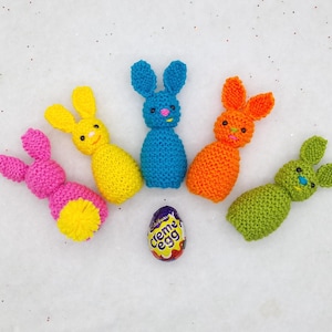 Könnte beinhalten: Fünf handgestrickte Osterhasen-Figuren in Pink, Gelb, Blau, Orange und Grün, angeordnet um ein Schokoladen-Creme-Ei. Die Häschen haben kleine, gestickte Gesichter und stehen vor einem weißen Hintergrund mit Glitzer.