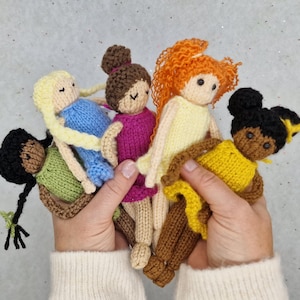 Puede incluir: Una colección de cinco muñecas tejidas a mano, cada una con características únicas. Las muñecas tienen diferentes tonos de piel, colores de cabello y atuendos en tonos de verde, azul, rosa, amarillo y marrón. Las muñecas miden unos 15 cm de alto.