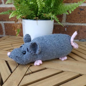 May include: A gray knitted mouse toy featuring a soft, textured body, rounded ears, and a small pink nose. The mouse has black bead eyes, thin black thread whiskers, and small pink feet with a matching pink tail extending from the back end.
