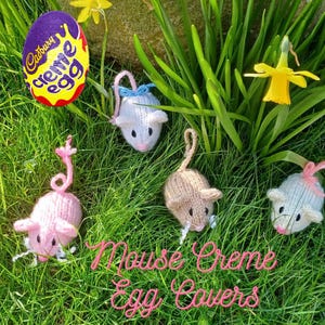 Puede incluir: Cuatro cubiertas de huevo de ratón de punto en rosa, beige y azul, sentadas en un lecho de hierba verde con narcisos amarillos. El texto "Mouse Creme Egg Covers" es visible en la imagen.