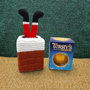 Könnte beinhalten: Ein gestrickter Schornstein-Überzug mit einem Paar rot-schwarzer Beine, die oben herausragen, neben einer blauen Schachtel Terry's Chocolate Orange. Der Schornstein hat einen weißen oberen Teil und eine braune Ziegelmuster-Basis.