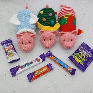 May include: Three knitted pig toys dressed as an angel, a Christmas tree, and an elf, with various chocolate bars in the background.