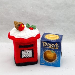 May include: A red knitted post box cover featuring a white snow-like top, a small brown robin with a red breast, and green holly leaves with red berries. The front displays a white square with the black text XMAS POST. Beside it is a Terry's Chocolate Orange box.