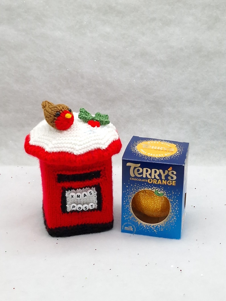 May include: A red knitted post box cover featuring a white snow-like top, a small brown robin with a red breast, and green holly leaves with red berries. The front displays a white square with the black text XMAS POST. Beside it is a Terry's Chocolate Orange box.