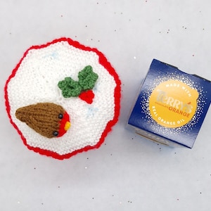 May include: A round, white crocheted cover with a red scalloped border, featuring a small brown robin bird with a red chest and green holly leaves with a red berry. Next to it is a blue square box labeled Terry's Chocolate Orange.