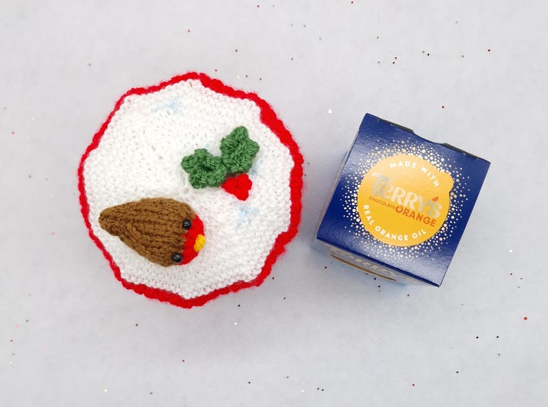 May include: A round, white crocheted cover with a red scalloped border, featuring a small brown robin bird with a red chest and green holly leaves with a red berry. Next to it is a blue square box labeled Terry's Chocolate Orange.