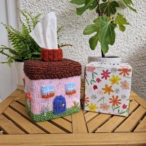 Puede incluir: Una funda para caja de pañuelos tejida con forma de pequeña casa con fachada rosa, techo marrón oscuro, puerta azul y dos ventanas. La funda presenta césped verde en la base y detalles florales. Al lado hay una caja blanca con un estampado floral colorido.