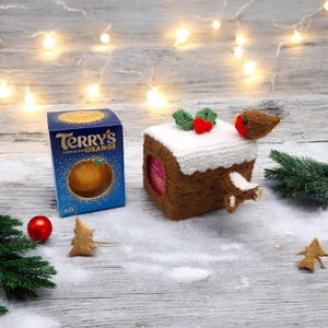 May include: A blue Terry's Chocolate Orange box next to a brown crocheted cozy featuring a white top, green leaves, red berries, and a small brown bird. The cozy is designed to fit around a round chocolate orange, with a cutout showing the packaging.