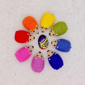 May include: A circle of colourful knitted hedgehogs surrounding a Cadbury Creme Egg. The hedgehogs are in the colours of the rainbow, starting with orange and ending with blue.