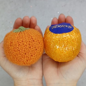 May include: Two orange-colored objects, one is a knitted orange and the other is a chocolate orange wrapped in gold foil with the text "Terry's" on the top.