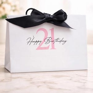 May include: A white rectangular gift bag featuring a large light pink number 21 printed on the front, overlaid with the black cursive text Happy Birthday. The bag is finished with a black satin ribbon bow tied at the top centre handle area.