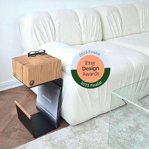 Könnte beinhalten: Ein moderner Beistelltisch mit einer Tischplatte aus Naturholz und einem Zeitschriftenständer aus schwarzem Metall. Der Tisch ist an einem weißen Sofa befestigt. Eine Brille liegt auf dem Tisch. Der Tisch ist Finalist der Etsy Design Awards 2023.
