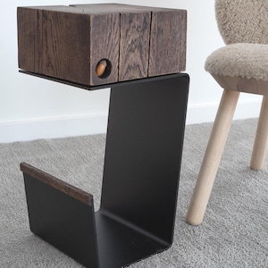 Op de afbeelding: Een moderne bijzettafel met een donkerhouten blokblad en een zwarte metalen basis. Het houten blok heeft een uitgesneden eikel detail. De tafel is ontworpen met een uitkragend blad en een tijdschriftenrek.