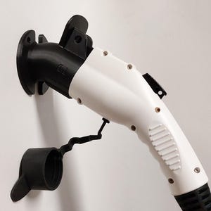 Op de afbeelding: Een wit en zwarte elektrische voertuig oplaadconnector. De connector heeft een afgeronde witte behuizing met een zwarte handgreep en een zwarte dop. De connector is op een witte muur gemonteerd.
